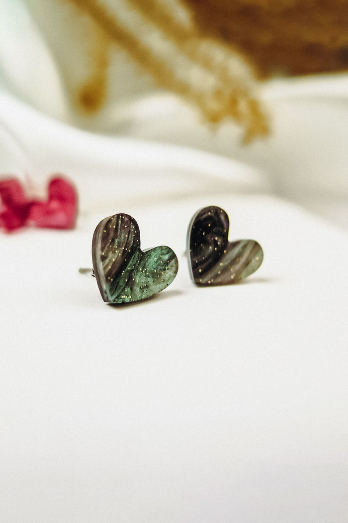 Two Tiny Heart Studs with a marbled green and black pattern are displayed on a white surface, exuding minimalist charm. Perfect for sensitive ears, these hypoallergenic earrings add a subtle touch of style amid softly blurred decor.