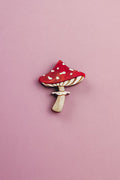 A handcrafted, eco-friendly Toadstool Mushroom Pin, shaped and painted like a red and white mushroom, rests on a light pink background.