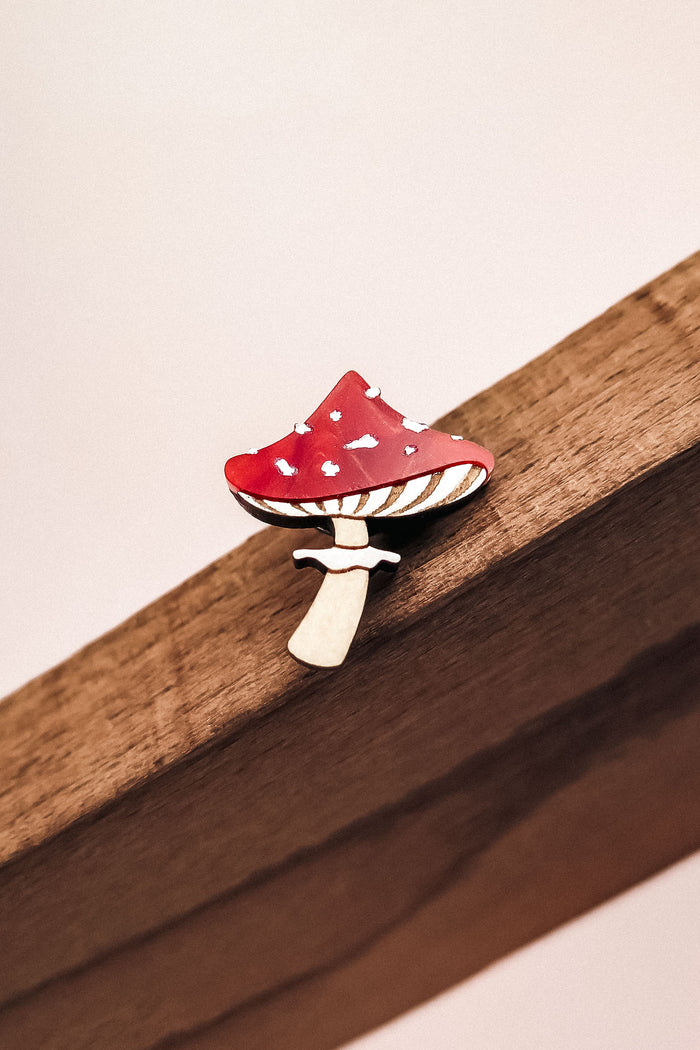 A small, handcrafted Toadstool Mushroom Pin shaped like a red and white spotted mushroom is attached to a wooden surface.