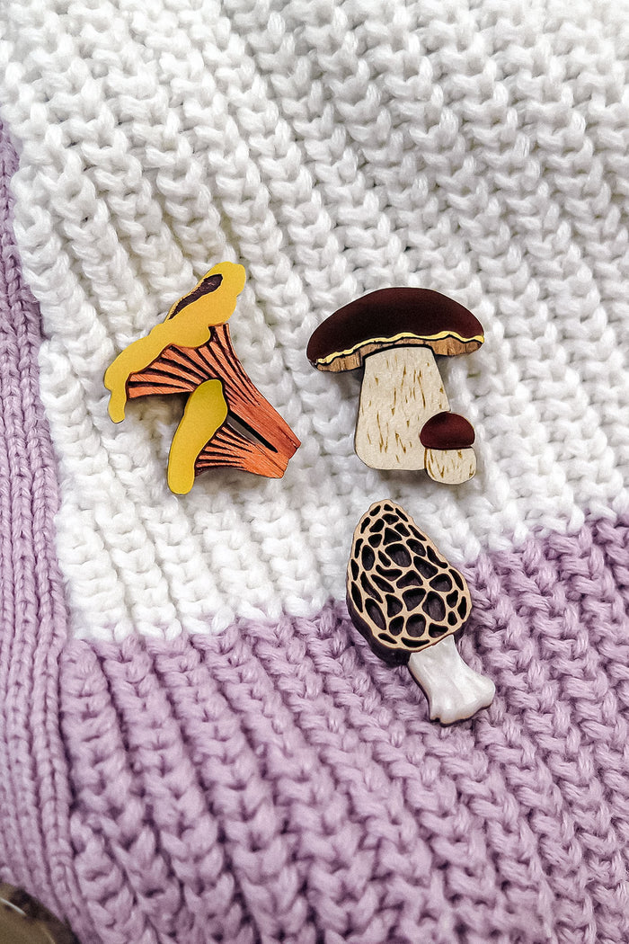 Three decorative mushroom pins, handcrafted from eco-friendly materials—including a Morel Mushroom Pin with a honeycomb pattern—are displayed on a knitted blanket with white and lavender stripes.