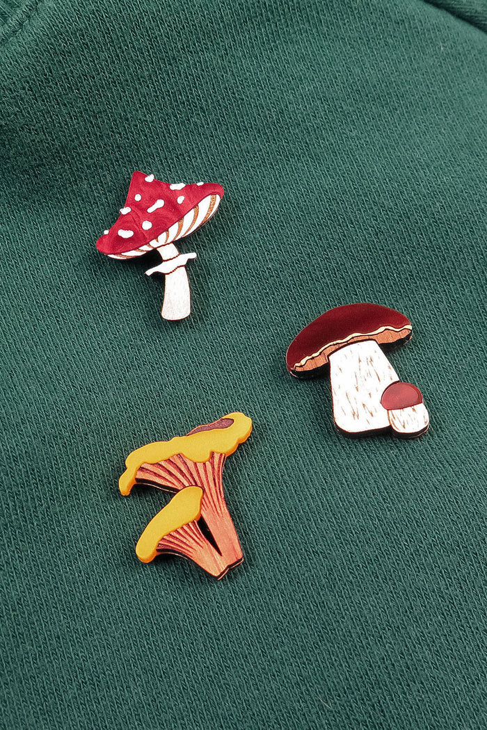 Three colorful, handcrafted enamel pins shaped like different mushrooms—including a Morel Mushroom Pin—are attached to a green fabric surface. These vibrant pins are made using eco-friendly materials for a sustainable touch.