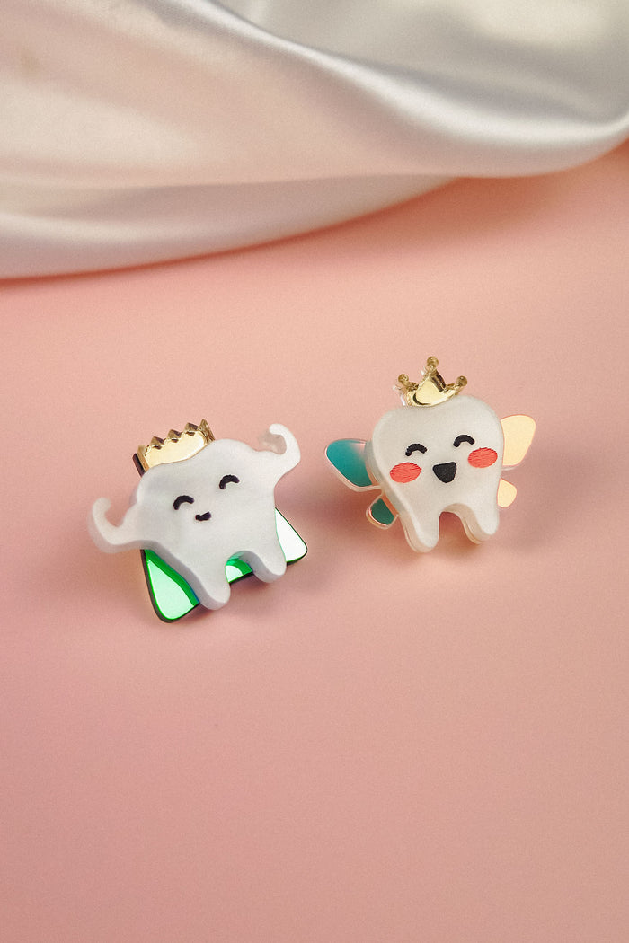 Two cute enamel pins shaped like smiling cartoon teeth, each a handcrafted accessory and true tooth king pin. One wears green wings, the other blue and gold with rosy cheeks, both crowned in gold against a soft pink background.