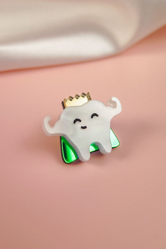 A cute handcrafted accessory, this enamel Tooth King Pin features a smiling tooth with little arms, a gold crown, and a shiny green base against a pink background with a white cloth in the corner.