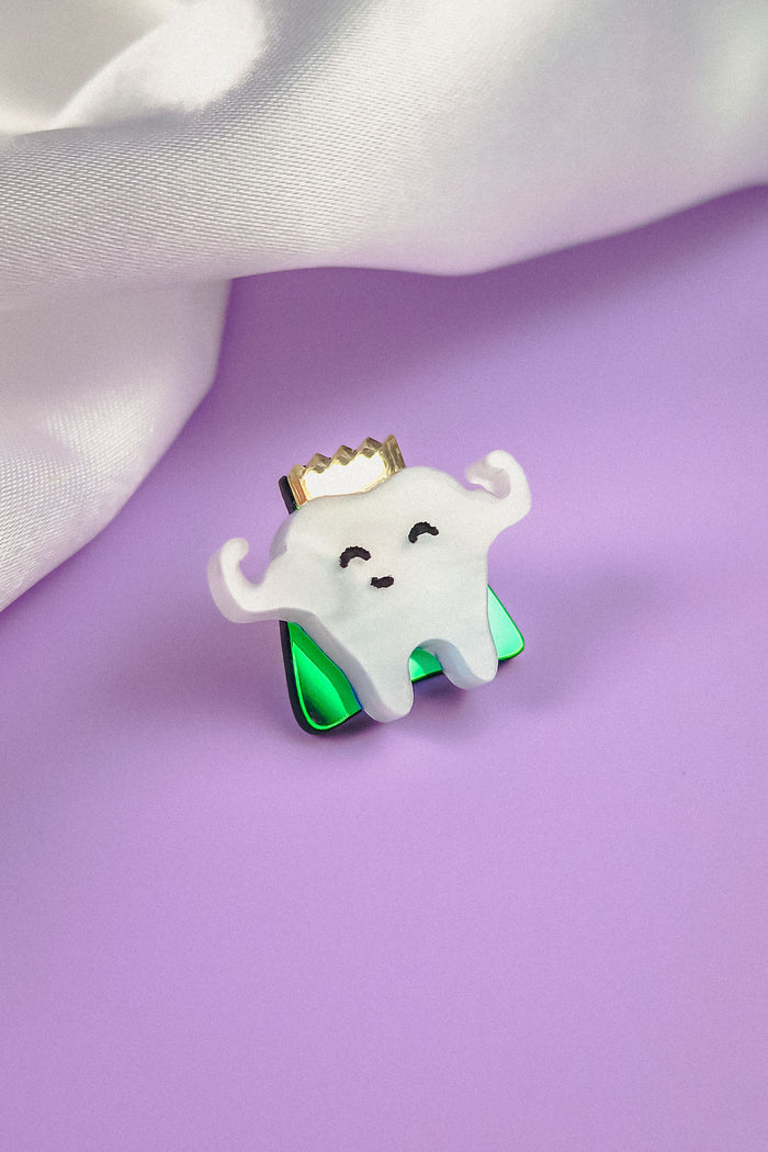 A cute Tooth King Pin shaped like a smiling tooth with a gold crown and flexed arms sits on a shiny green base, placed on a purple surface with white fabric in the background—a charming handcrafted accessory for any collection.