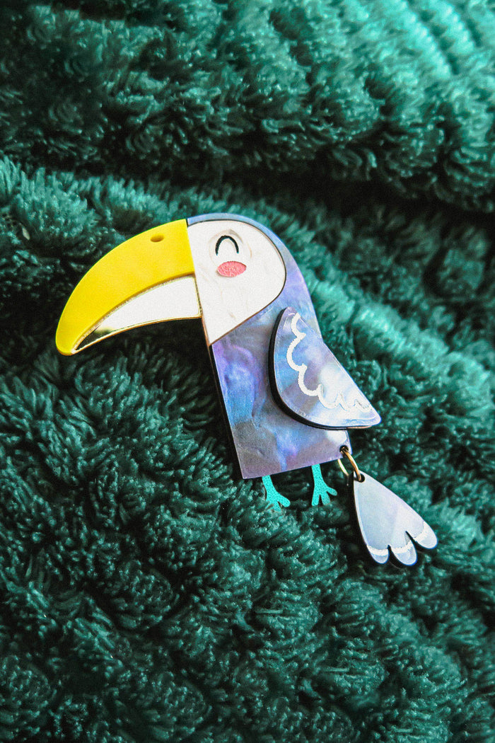 A handcrafted Toucan Brooch with a yellow beak and smiling face brings tropical charm as it rests on a textured dark green fabric background.