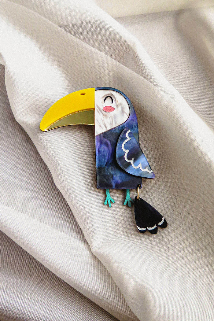 A handcrafted Toucan Brooch, featuring a bright yellow beak and a dangling tail, brings tropical charm as it rests on soft, cream-colored fabric.