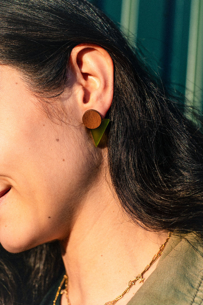 Close-up of a person with dark hair wearing striking triangle earrings crafted from acrylic glass, paired with a gold chain necklace. The background features blurred green vertical lines, adding to the modern aesthetic.