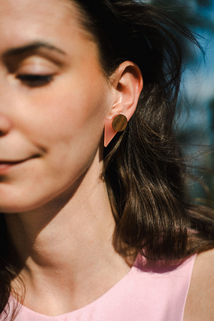 A woman with long brown hair wears striking triangle earrings made of acrylic glass, featuring pink and gold shapes. She is dressed in a light pink top and stands outdoors in sunlight, only the left side of her face visible with a soft smile.