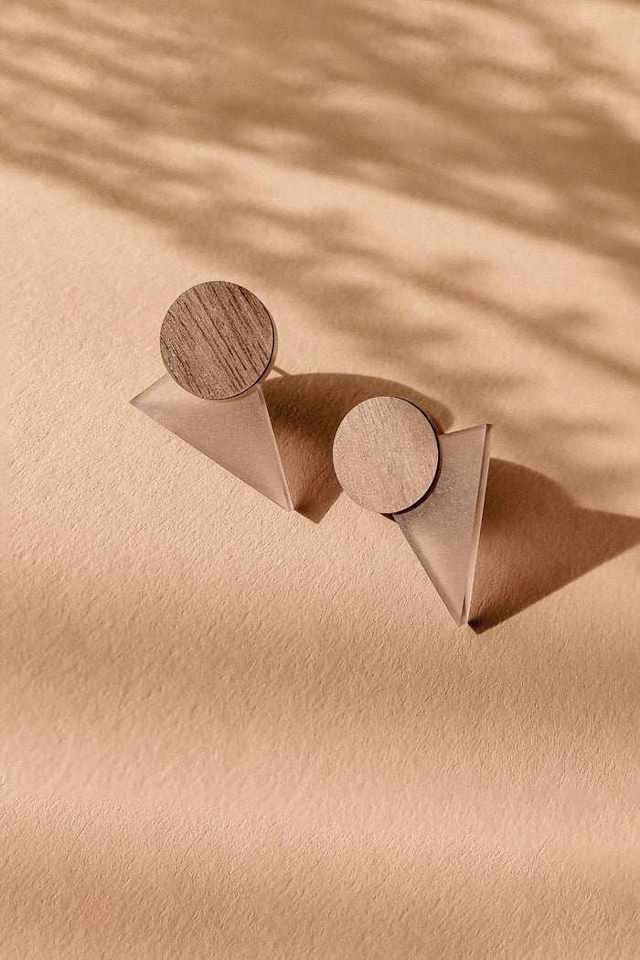 A pair of modern, handcrafted earrings featuring wooden circular tops and translucent triangular bases, displayed on a textured beige surface with soft shadows—perfect for a contemporary capsule wardrobe.