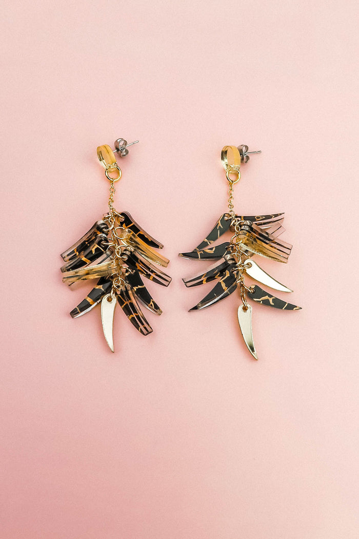 A pair of featherlight dangle earrings with layered gold and tortoiseshell-patterned pieces arranged in a fan-like design, displayed on a light pink background.