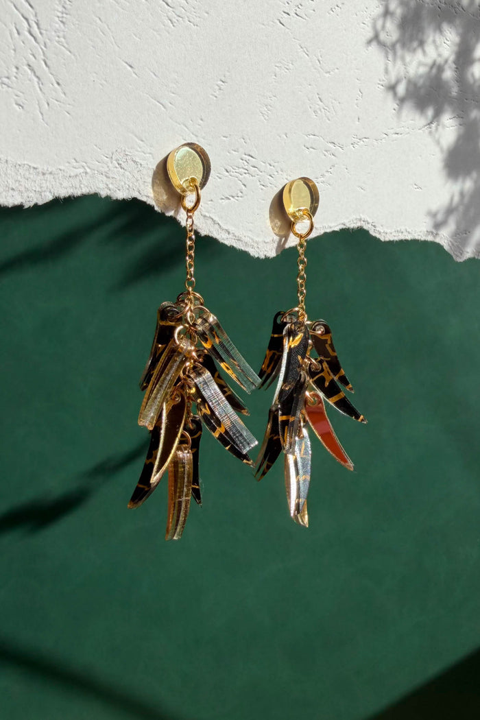 Featherlight gold and tortoiseshell dangle earrings with layered, leaf-like charms hang from a white textured surface above a green background. Light casts soft shadows on the wall.