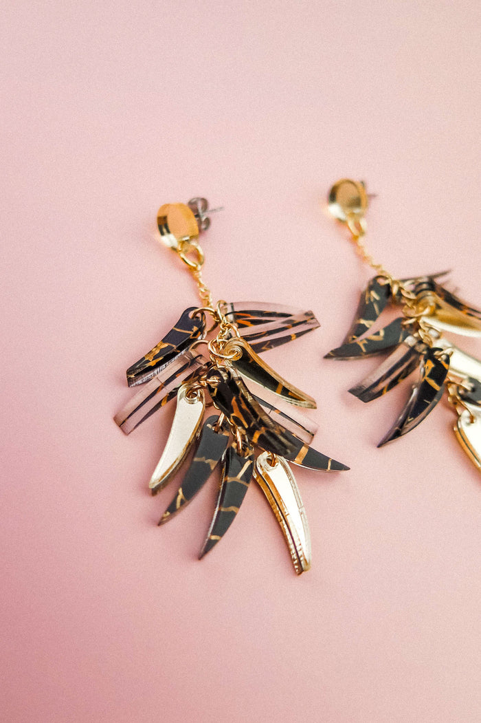 Gold and black marbled featherlight dangle earrings with multiple layered, leaf-shaped pendants arranged in a fan shape, displayed on a soft pink background.