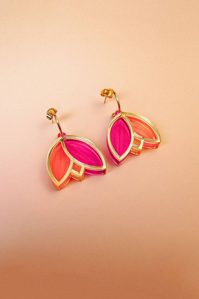 A pair of Tulip Huggie Earrings with artisan craftsmanship, featuring gold hoops and overlapping leaf-shaped charms in vibrant pink and orange colors, displayed on a peach-colored background.