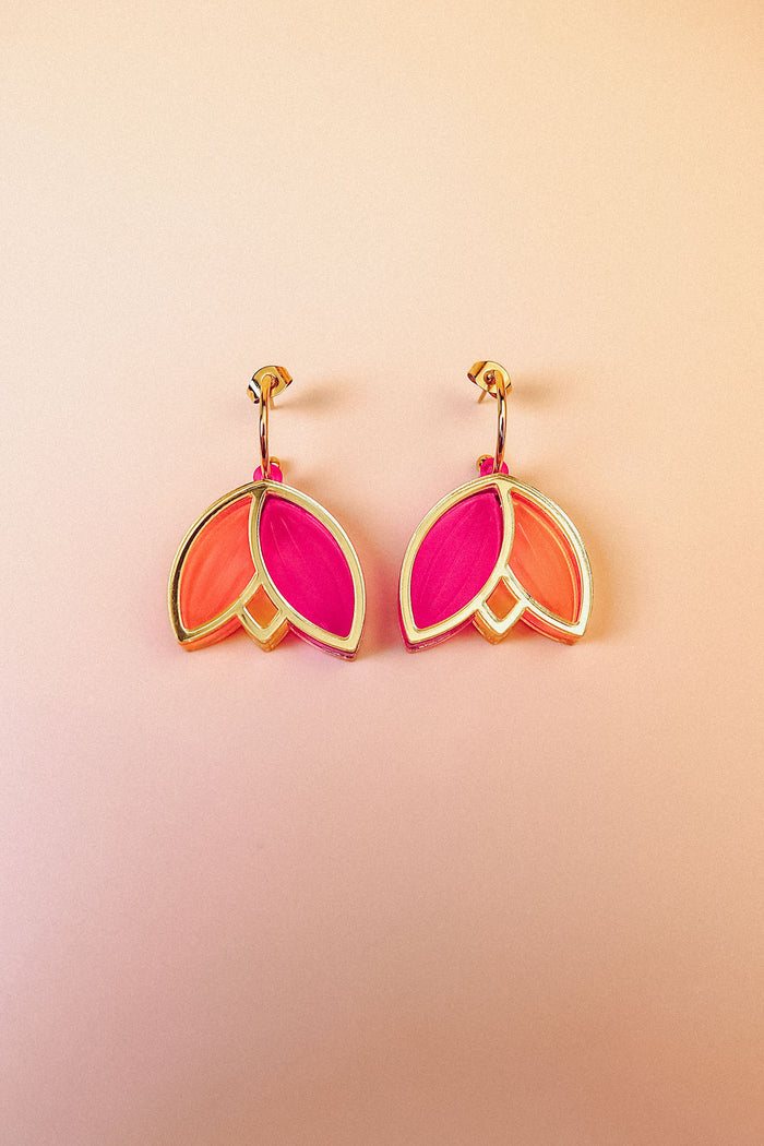 A pair of Tulip Huggie Earrings with three petal-shaped sections—two orange and one bright pink—arranged in a floral design, showcasing artisan craftsmanship on a light peach gradient background.