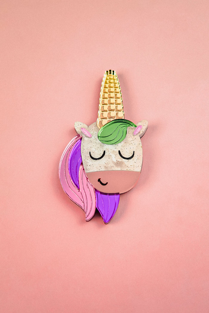 A playful accessory, this handcrafted UniCorn Brooch features a cute unicorn face with closed eyes, a golden horn, green mane, and pink and purple hair on a pastel pink background.