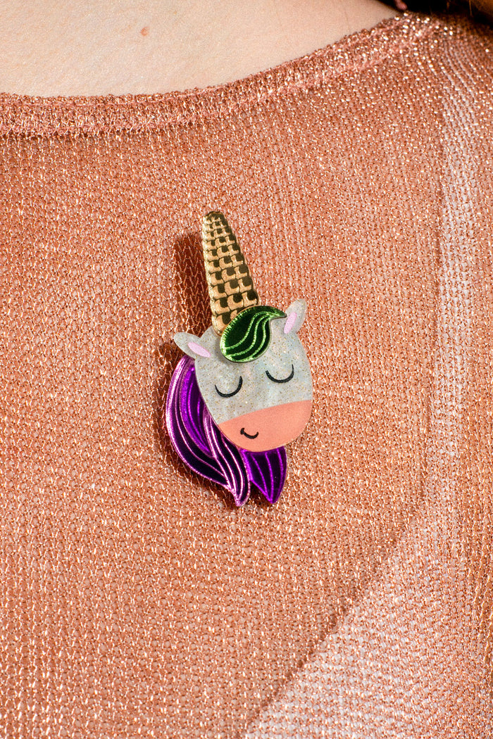 A glittery unicorn pin with a gold horn, green mane, and purple hair is attached to a pink shimmery fabric—making this UniCorn Brooch a delightful and playful accessory that adds a whimsical touch to any outfit.