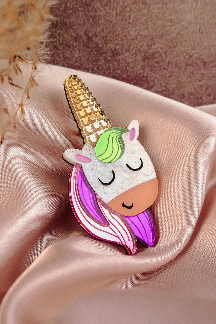 A cute handcrafted UniCorn Brooch with a gold horn, green mane, pink-and-purple hair, and closed eyes rests on pink satin fabric—a playful accessory for any outfit.
