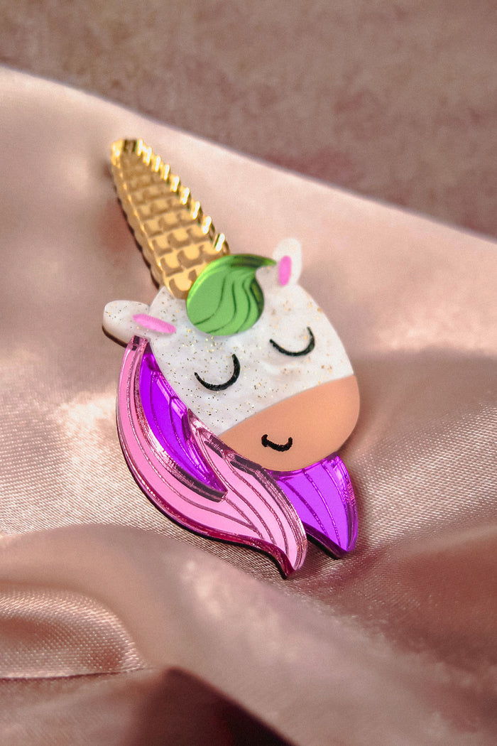 A colorful handcrafted UniCorn Brooch shaped like a unicorn’s head with a golden horn, green mane, closed eyes, and pink and purple details rests on shiny light pink satin fabric—a playful accessory for any collection.