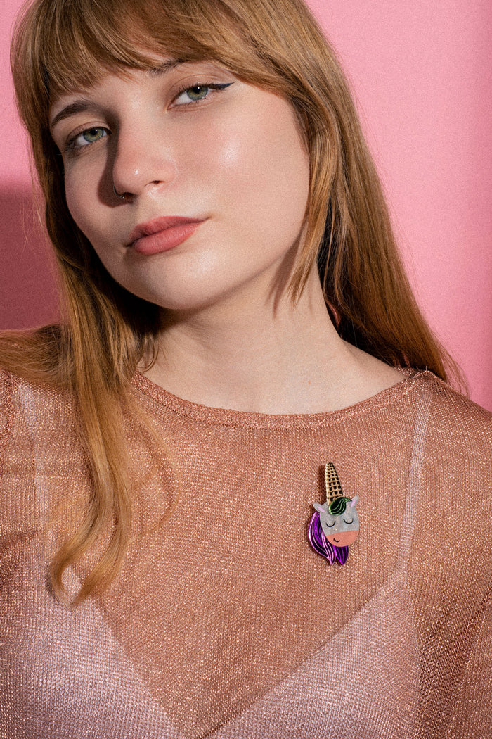 A woman with long blonde hair and winged eyeliner poses against a pink background, wearing a sheer, shimmery top adorned with a playful accessory—a handcrafted UniCorn Brooch featuring purple hair and a gold horn.