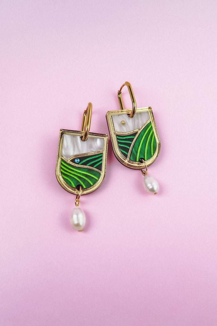 A pair of Vineyard Inspired Art Nouveau Dangle Earrings with green and white enamel landscape designs, accented with pearls & rhinestones and hanging pearl drops, displayed on a light pink background.