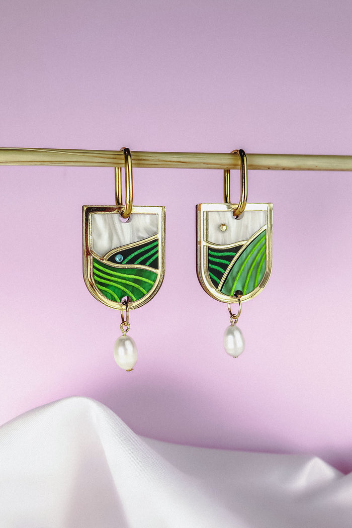 Gold Vineyard Inspired Art Nouveau Dangle Earrings with green and white enamel, an abstract landscape design, and small pearl drops, hang from a horizontal rod against a soft pink and white background.
