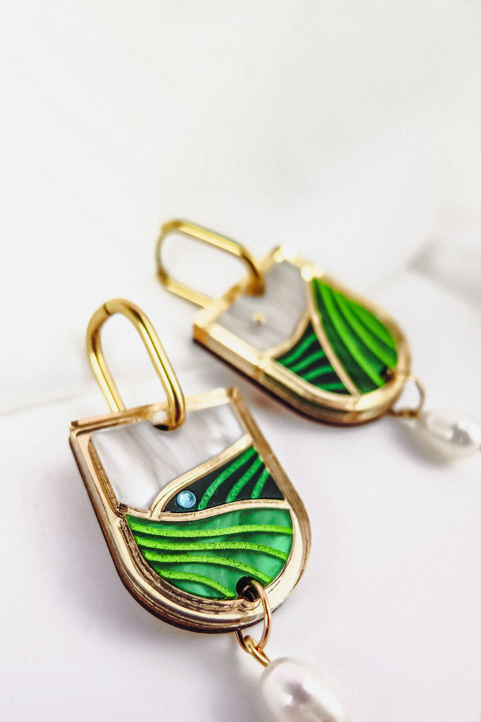 Close-up of Vineyard Inspired Art Nouveau Dangle Earrings, featuring green and white enamel designs, a touch of blue, and a dangling white pearl, displayed on a white surface.