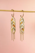 A pair of Wheat Statement Earrings with cut-out leaf patterns and dangling chain accents hang from a horizontal rod against a soft pink background, showcasing standout style.