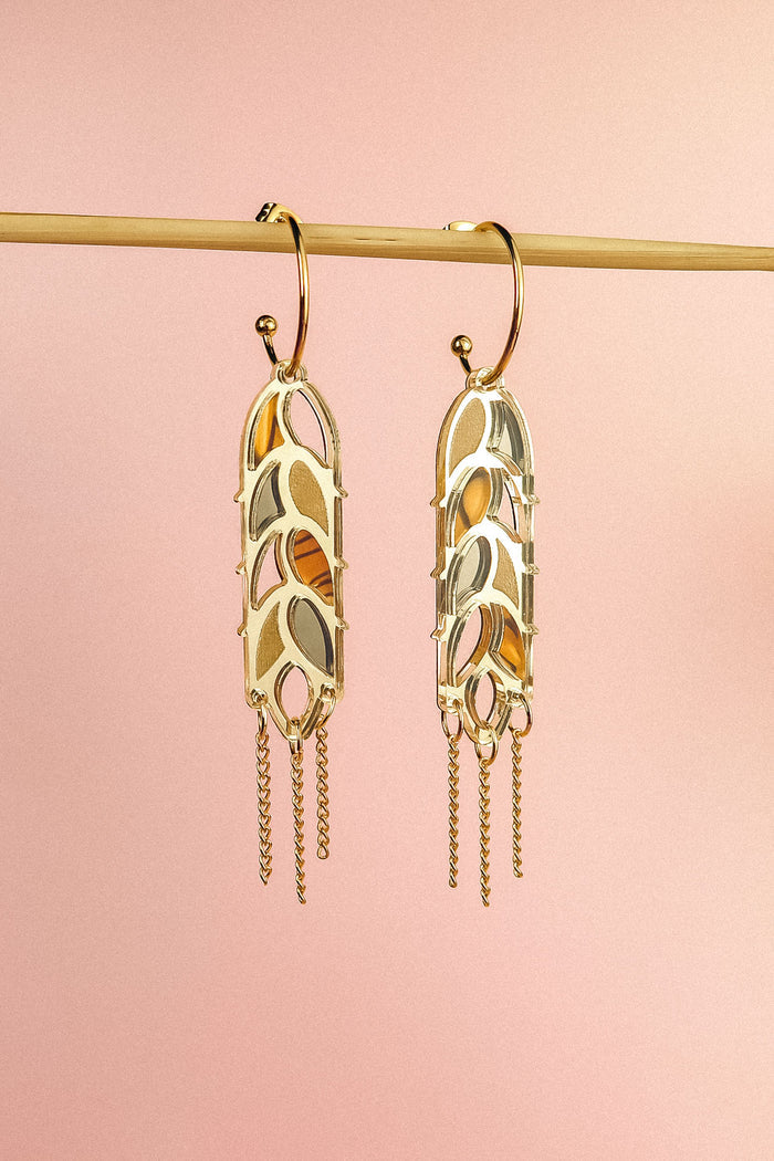 A pair of Wheat Statement Earrings with cut-out leaf patterns and dangling chain accents hang from a horizontal rod against a soft pink background, showcasing standout style.