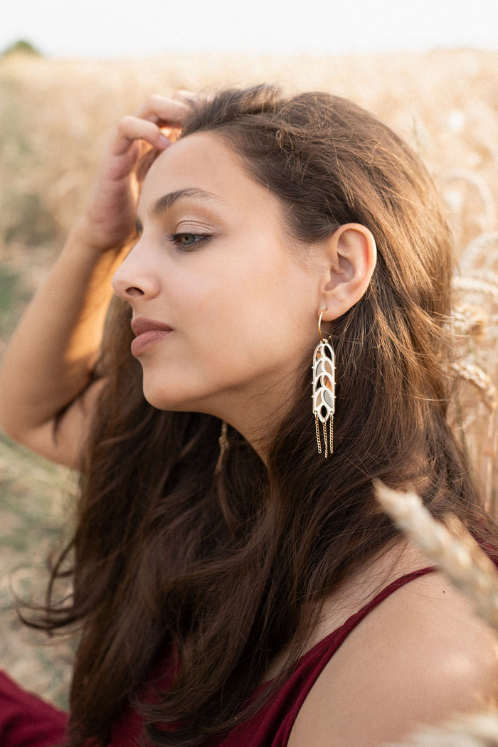 A young woman with long brown hair sits in a wheat field, wearing a burgundy dress and Wheat Statement Earrings. She gazes thoughtfully to the side, lightly touching her hair, her standout style enhanced by the bold design of her earrings.
