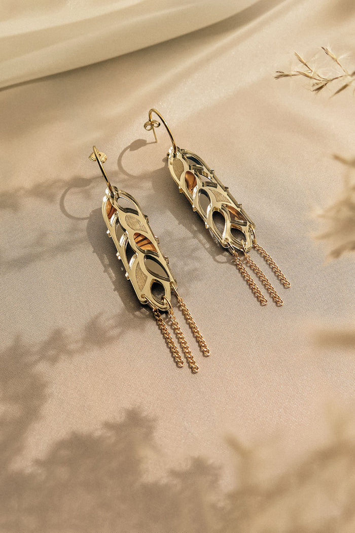 A pair of Wheat Statement Earrings with intricate cutout designs and dangling chain accents, displayed on a soft beige surface with delicate shadows and dried flowers—a bold design perfect for standout style.